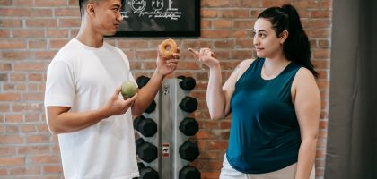 Side view of Asian personal instructor with apple and doughnut offering healthy food to plus size woman while standing in gym