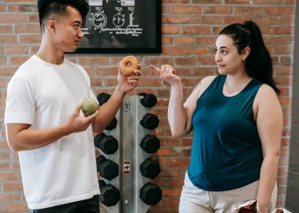 Side view of Asian personal instructor with apple and doughnut offering healthy food to plus size woman while standing in gym